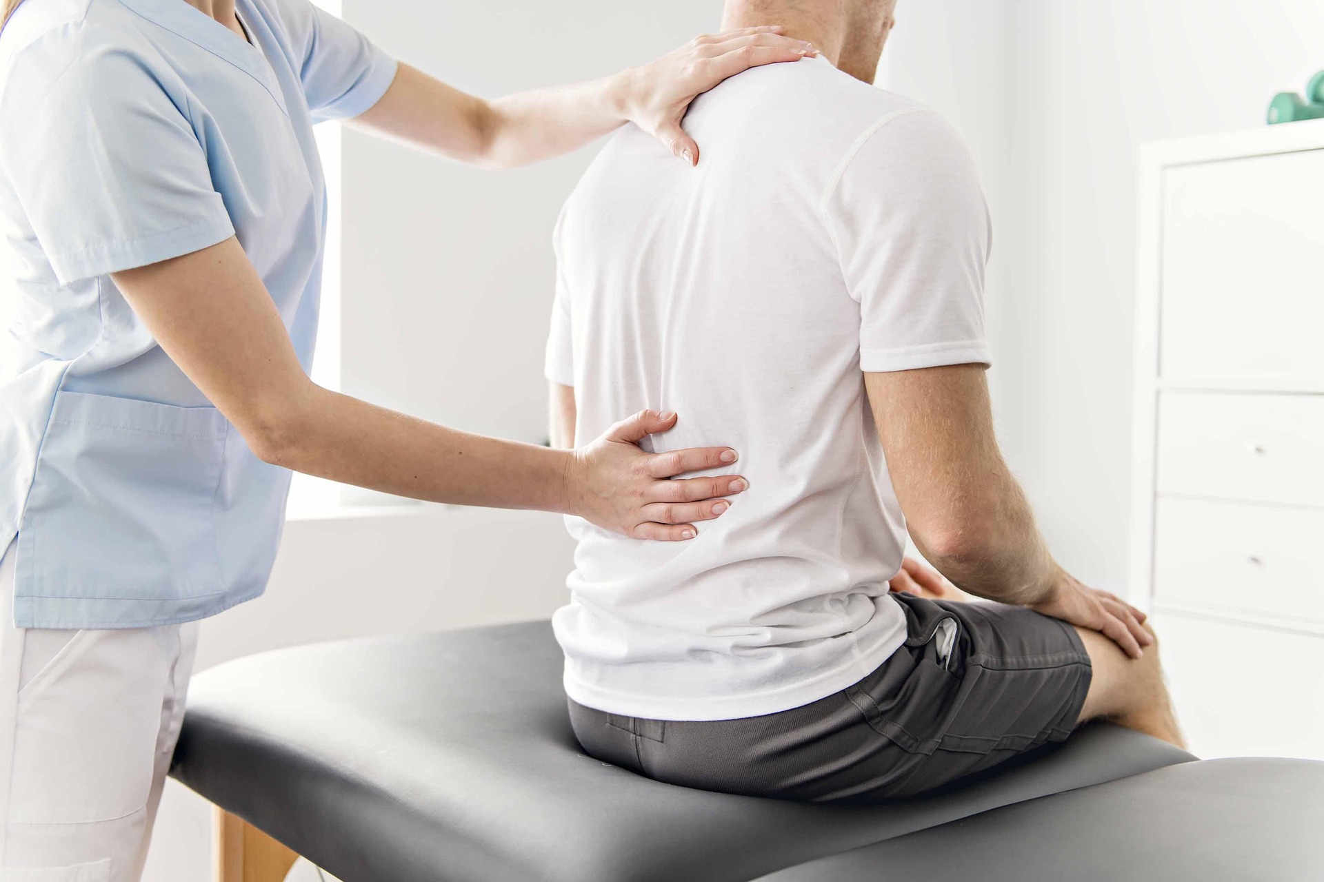 A patient wearing a white t shirt sitting on exam table while you see the practitioner hands on the patient's back while doing an exam